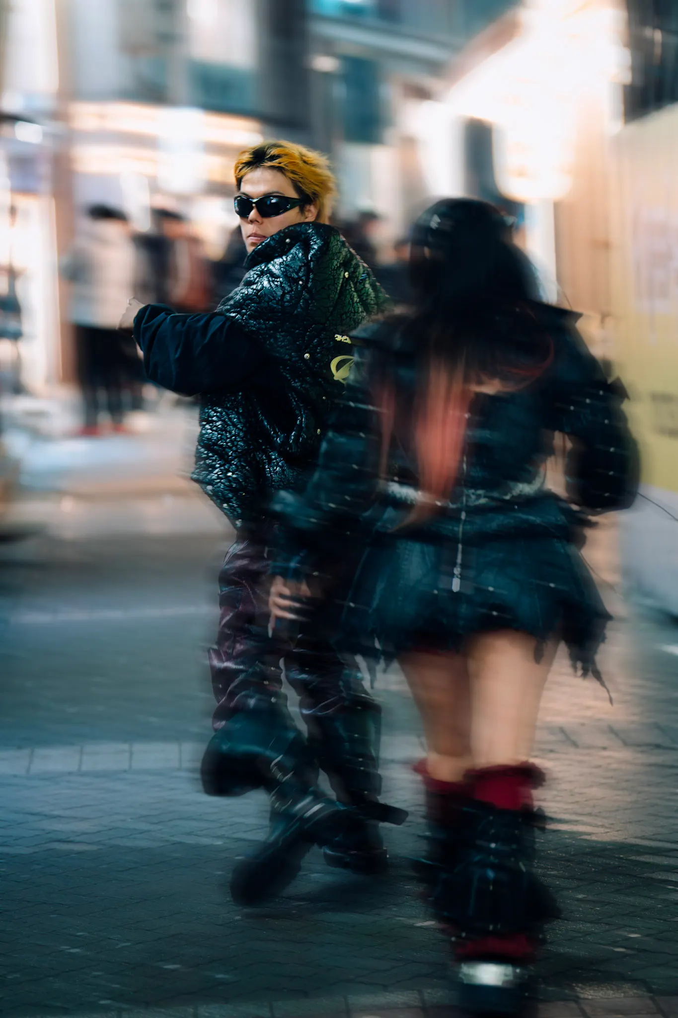 Motion-blurred night street scene: a person with dyed blond hair and sunglasses in a glossy black jacket walking past a blurred figure in a 